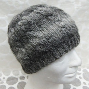 May include: A gray and white knitted beanie hat with a cable knit design. The hat has a ribbed brim and a rounded top. The color gradient goes from dark gray to light gray and white.