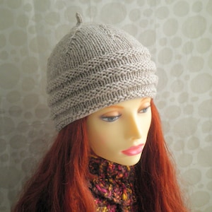 Puede incluir: Un gorro tejido de color beige claro con una pequeña parte superior puntiaguda. El gorro tiene un borde acanalado y un patrón de tejido texturizado. El gorro se muestra en un maniquí con cabello rojo y una bufanda colorida.