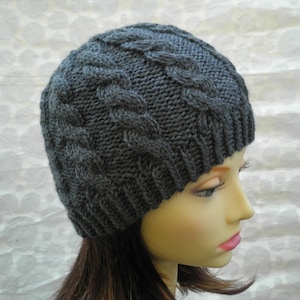 May include: A dark gray knitted beanie hat with a cable knit design. The hat is made of a soft, textured yarn and has a ribbed brim. The cable knit pattern covers the top of the hat. The hat is displayed on a mannequin head.