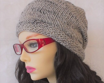 May include: A gray knitted beanie with a black fleck pattern. The beanie is worn by a person with long dark hair and red-framed glasses.
