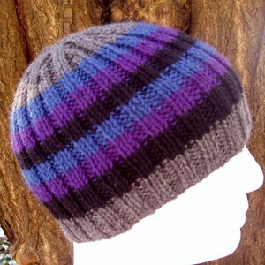 Op de afbeelding: Een gebreide beanie met horizontale strepen in tinten grijs, blauw, paars en zwart. De hoed is gemaakt van een zacht, getextureerd garen en heeft een geribbelde rand. De hoed is ontworpen om nauwsluitend op het hoofd te passen.