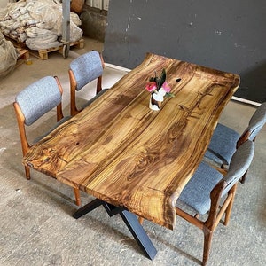 Puede incluir: Una mesa grande de madera de borde vivo con un acabado de madera natural. La mesa tiene una base de metal negro y está rodeada por cuatro sillas tapizadas grises. Un pequeño jarrón con flores rosas se encuentra sobre la mesa.