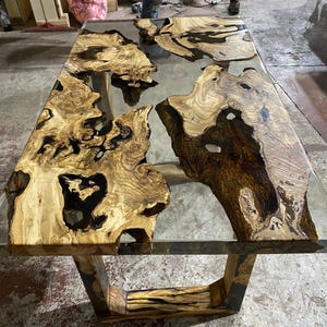 May include: A rectangular wooden table with a clear epoxy resin top. The wood has a natural grain pattern in shades of brown and tan. The table is supported by a wooden base.