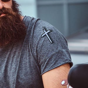 May include: A charcoal gray short-sleeve t-shirt featuring a small, embroidered white cross patch on the left shoulder. The shirt has a crew neckline and rolled sleeve cuffs, crafted from a heathered fabric with a textured appearance.