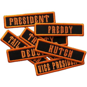 May include: A collection of black rectangular patches with orange embroidered text and borders. The patches display various titles and names, including "PRESIDENT", "FREDDY", "HUTCH", and "VICE PRESIDENT". The patches are likely for clothing or accessories.