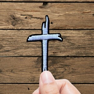 May include: An embroidered patch in the shape of a cross with a light blue thread finish and a black border. The cross features a textured, slightly uneven design with frayed-style edges, creating a distinct outline against the background.