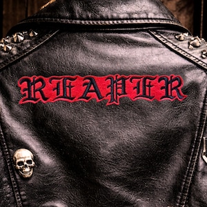 May include: Black leather jacket with the word "REAPER" embroidered in red and navy blue gothic lettering across the back. The jacket features silver-colored studs on the shoulders and a skull pin.