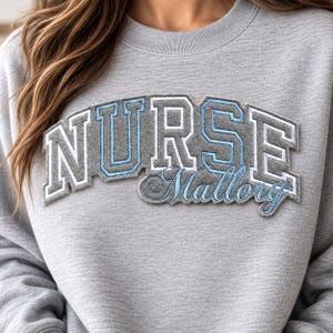 May include: A light grey crewneck sweatshirt featuring an arched applique patch with the word NURSE in grey felt with white and light blue stitched borders. Below, the name Mallory is embroidered in light blue cursive script lettering.