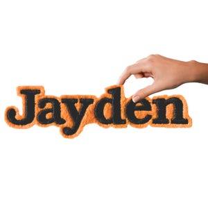 May include: A custom name patch featuring the name Jayden in black, textured, fuzzy lettering. The letters are outlined with a thick, vibrant orange, fuzzy border, creating a bold contrast between the dark text and the bright background.