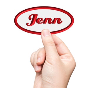 Pode incluir: Uma mão segura um adesivo oval branco com uma borda vermelha. O adesivo diz "Jenn" em letras vermelhas e cursivas.