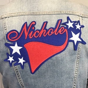 May include: A blue denim jacket with a red, white, and blue patch on the back. The patch features the name "Nichole" in blue lettering with white stars.