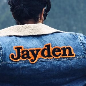 May include: A blue denim jacket featuring a cream-coloured sherpa collar and a large, orange and black felt patch on the back with the name Jayden stitched in a bold, rounded font. The jacket includes metal buttons along the bottom hem.