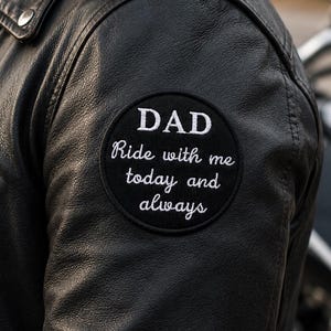 Può includere: Una giacca in pelle nera con una toppa circolare ricamata sulla manica. La toppa è nera con testo bianco che recita DAD Ride with me today and always. La giacca presenta una cerniera in metallo argentato e un bottone a pressione sul colletto.