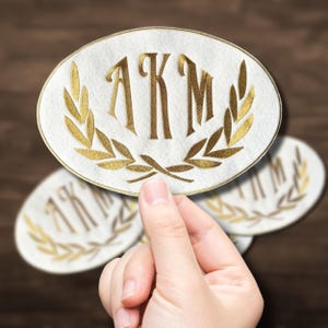 Peut inclure: Écusson ovale blanc avec les initiales "AKM" brodées en or et un motif de couronne de laurier doré. L'écusson est tenu dans une main, avec deux autres écussons légèrement flous en arrière-plan. Les écussons sont probablement destinés aux vêtements ou aux accessoires.