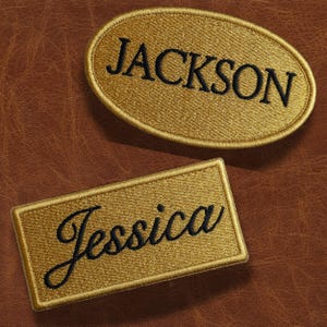 May include: Two embroidered patches with names. One is an oval shape with the name "JACKSON" in black thread. The other is a rectangle with the name "Jessica" in a cursive font. Both patches are gold with a brown background.