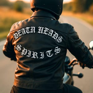 May include: Black leather motorcycle jacket featuring the words "DEATH HEADS SPIRITS" embroidered in white gothic lettering. The jacket is worn by a person riding a motorcycle on a road, with a black helmet.