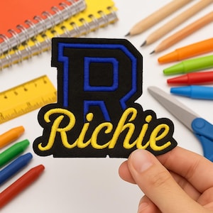 Peut inclure: Écusson brodé noir et bleu en forme de lettre « R » avec le nom « Richie » en jaune. L'image comprend des fournitures scolaires telles que des crayons, des stylos et une règle.