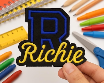 以下が含まれることがあります： 上部に大きな青い文字R、その下に黄色の筆記体でRichieという名前が刺繍された黒いワッペン。ワッペンには黒い縁取りがあり、質感のある生地で作られており、詳細なステッチが施されています。