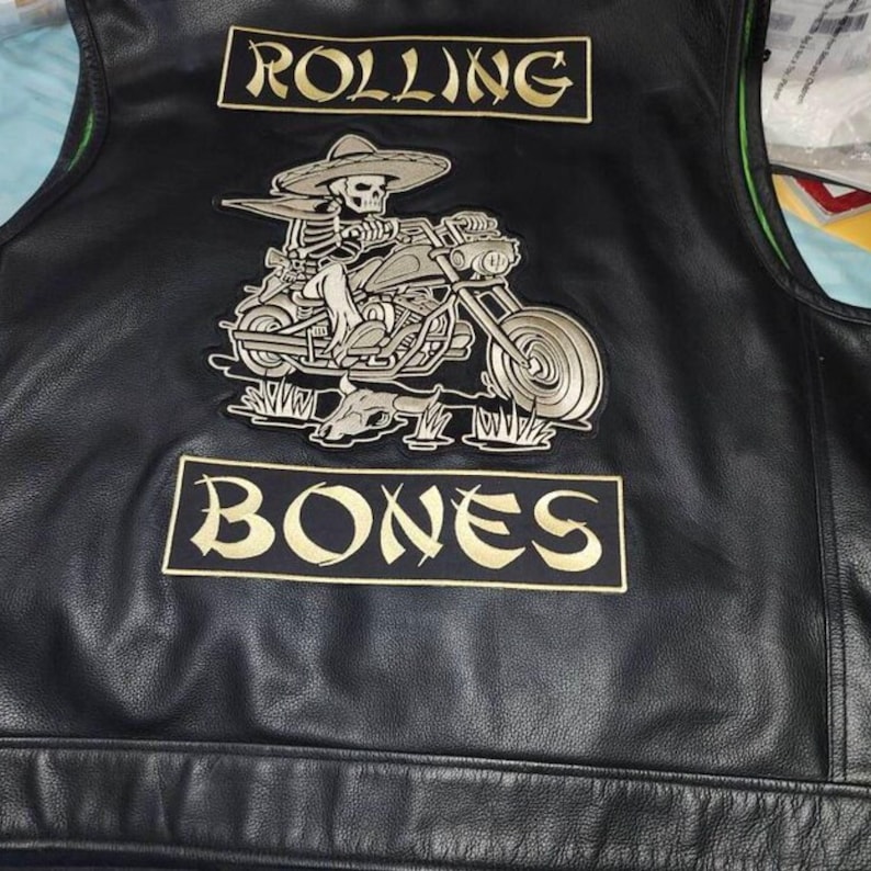 Puede incluir: Chaleco de cuero negro con un parche grande que presenta un esqueleto montando una motocicleta, con un sombrero. El texto "ROLLING" est&aacute; encima del parche y "BONES" debajo, ambos en letras doradas.