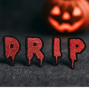 May include: Red embroidered patches spelling "DRIP" with a black border, featuring a dripping effect. A carved orange pumpkin is in the background. The patches are on a dark gray textured surface. Halloween theme.