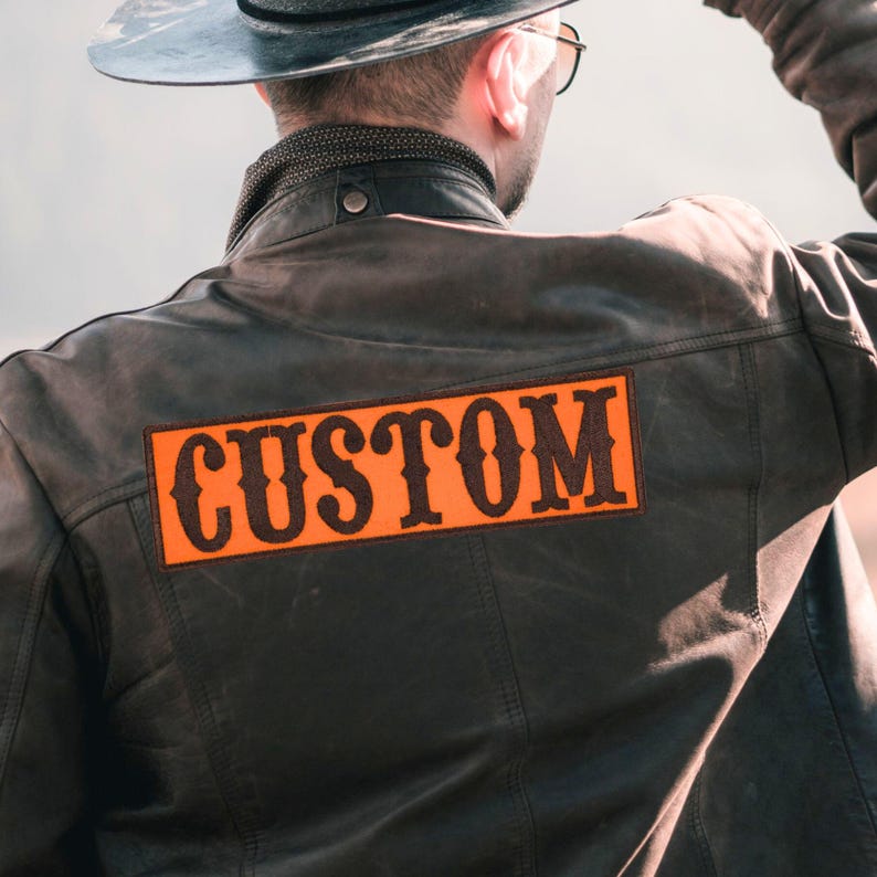 K&ouml;nnte beinhalten: Dunkelbraune Lederjacke mit dem Wort "CUSTOM" in orangefarbenen und schwarzen Buchstaben. Die Jacke hat ein klassisches Design mit sichtbaren N&auml;hten und einem abgenutzten Aussehen. Das Bild konzentriert sich auf die R&uuml;ckseite der Jacke und hebt den Aufn&auml;her hervor.