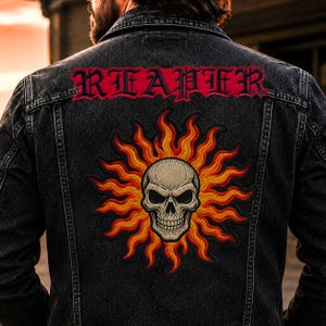 May include: A black denim jacket featuring a large embroidered patch on the back. The patch displays a beige skull centered within a vibrant sunburst of orange and red flames. Above the skull, the word REAPER is embroidered in red gothic font.