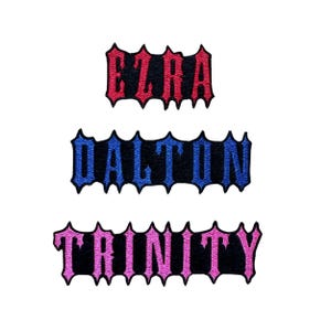 Puede incluir: Tres parches de tela bordados con bordes dentados de estilo g&oacute;tico. El parche superior presenta el nombre EZRA en hilo rojo, el parche central muestra DALTON en hilo azul y el parche inferior muestra TRINITY en hilo rosa sobre tela negra.