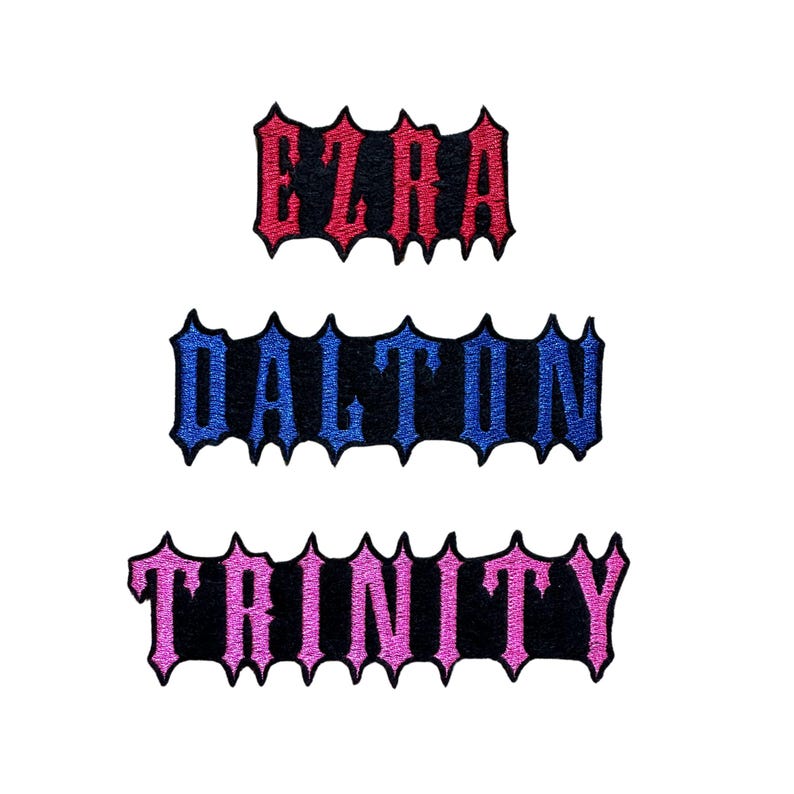Puede incluir: Tres parches de tela bordados con bordes dentados de estilo g&oacute;tico. El parche superior presenta el nombre EZRA en hilo rojo, el parche central muestra DALTON en hilo azul y el parche inferior muestra TRINITY en hilo rosa sobre tela negra.