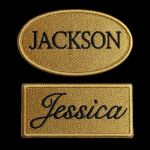 Op de afbeelding: Twee gouden, geborduurde naamlabels. Het bovenste label is ovaal en leest "JACKSON" in zwarte letters. Het onderste label is rechthoekig en toont de naam "Jessica" in een cursief lettertype. Beide labels zijn goudkleurig.