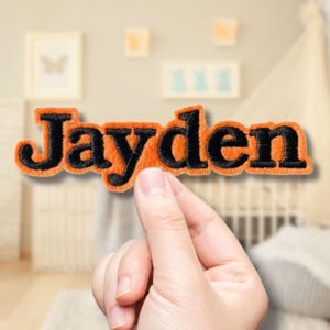 May include: An orange and black embroidered patch with the name "Jayden". The letters are black with an orange border. The patch is held up by a hand in front of a blurred background.