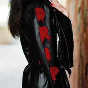 May include: Black leather jacket featuring the word "DRIP" in red, dripping-style lettering on the sleeve. The jacket has a zip closure and a belted waist. The overall style is edgy and modern.
