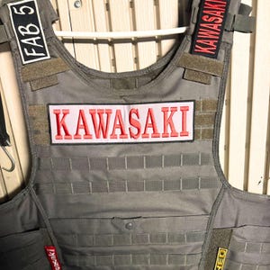 Puede incluir: Un chaleco t&aacute;ctico gris con m&uacute;ltiples parches. El parche central dice "KAWASAKI" en letras rojas sobre un fondo rosa. Parches adicionales incluyen "KAWASAKI" en los hombros. El chaleco tiene un patr&oacute;n de rejilla.