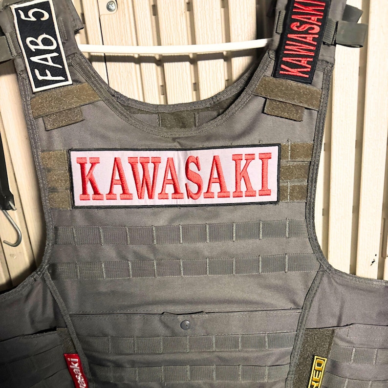 Puede incluir: Un chaleco t&aacute;ctico gris con m&uacute;ltiples parches. El parche central dice "KAWASAKI" en letras rojas sobre un fondo rosa. Parches adicionales incluyen "KAWASAKI" en los hombros. El chaleco tiene un patr&oacute;n de rejilla.