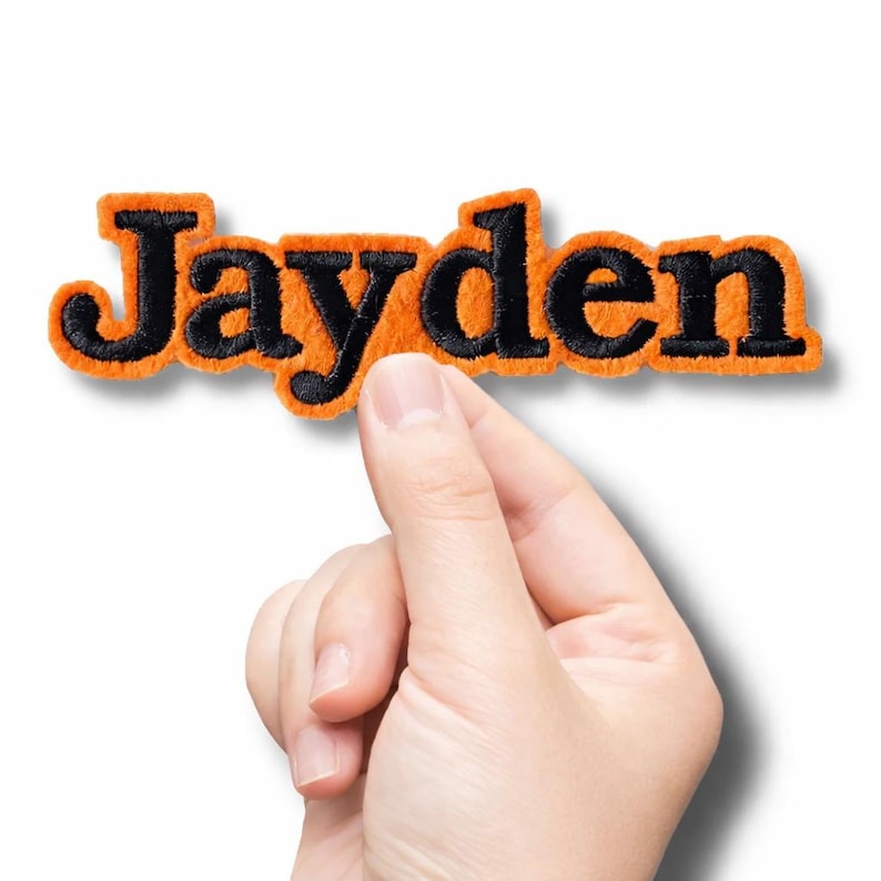 Puede incluir: Un parche con nombre personalizado que presenta el texto Jayden en letras bordadas negras con un borde naranja grueso y difuso. El parche tiene una apariencia suave y texturizada, dise&ntilde;ado para aplicarse sobre telas como elemento decorativo.