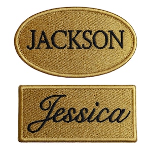 Puede incluir: Dos parches bordados con nombres. Uno es de forma ovalada con el nombre "JACKSON" en escritura negra. El otro es rectangular con el nombre "Jessica" en escritura negra. Ambos parches tienen un fondo dorado.