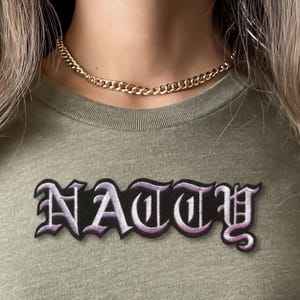 Puede incluir: Una camiseta verde oliva con un parche bordado negro que muestra la palabra NATTY en una fuente de estilo g&oacute;tico blanca y morada. Un collar de cadena de metal dorado con eslabones entrelazados descansa alrededor del cuello de la prenda.
