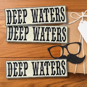 Puede incluir: Tres parches rectangulares con el texto "DEEP WATERS" en letras negras adornadas. Un recorte de bigote y gafas negras est&aacute; al lado de un regalo envuelto sobre una superficie de madera.