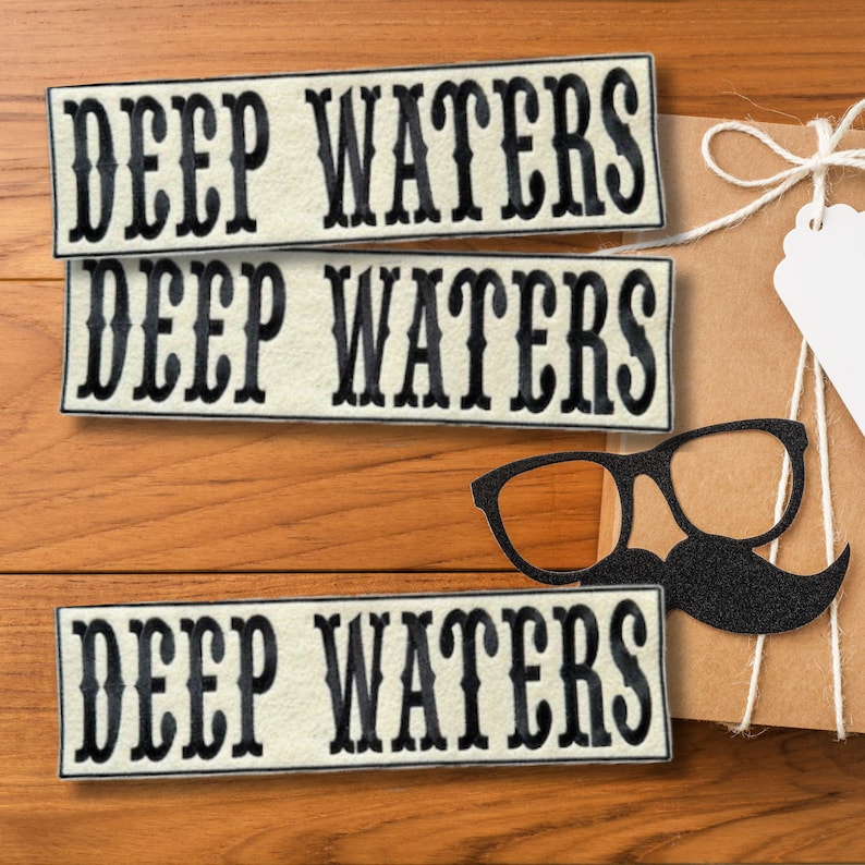 Puede incluir: Tres parches rectangulares con el texto "DEEP WATERS" en letras negras adornadas. Un recorte de bigote y gafas negras est&aacute; al lado de un regalo envuelto sobre una superficie de madera.