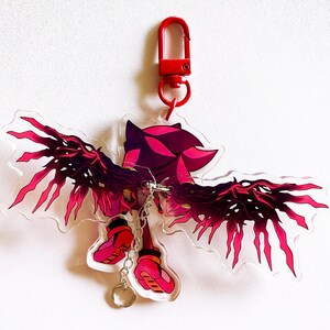 May include: A clear acrylic keychain featuring a stylized character with red and pink accents, including large wings and boots. The keychain has a red clasp and a silver keyring.