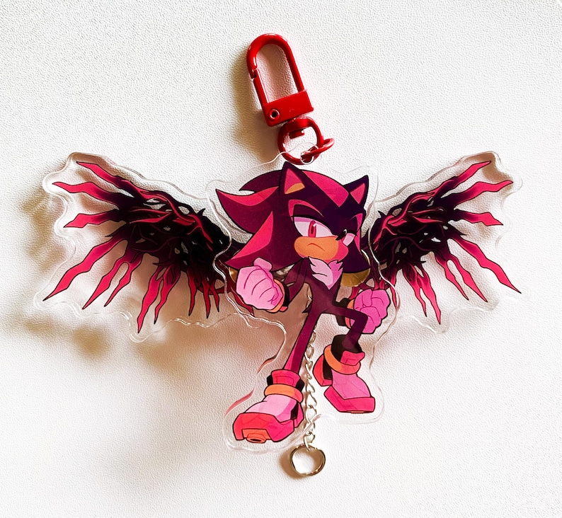 May include: A clear acrylic keychain featuring a character with red and black wings, a red clip, and a silver chain. The character is primarily red and black with pink accents. The keychain is designed to be attached to keys or a bag.