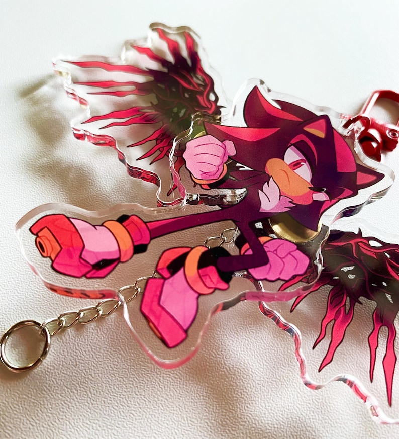 May include: A clear acrylic keychain featuring a cartoon character with a red and black color scheme. The character is in a dynamic pose, with pink and orange accents. The keychain includes a silver chain and a red clasp.