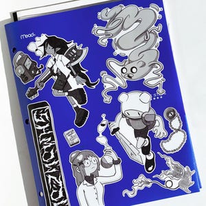 May include: A blue Mead binder covered in cartoon stickers. The stickers feature black and white illustrations of characters and objects. One sticker has the text "ZOMBIE". Other stickers include a character with an axe and a character holding a sword.