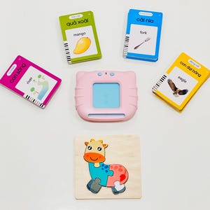May include: A collection of educational toys, including flashcards with images of a mango, fork, eagle, and river. A pink electronic reader and a wooden cow puzzle are also present. The flashcards have text in Vietnamese and English.