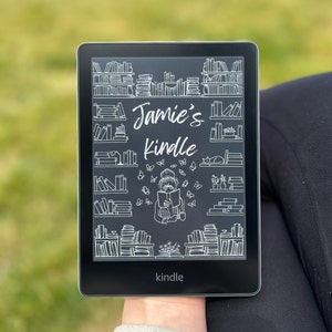 Puede incluir: Una ilustración en blanco y negro de una persona leyendo un libro en un lector electrónico Kindle. La ilustración está rodeada de estanterías llenas de libros. El texto "Jamie's Kindle" está escrito en la pantalla.