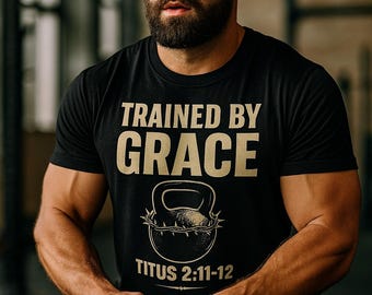 Puede incluir: Camiseta negra con el texto "TRAINED BY GRACE" y un diseño de pesa rusa. El texto "TITUS 2:11-12" está debajo. La persona sostiene una pesa rusa, lo que sugiere un tema de fitness o entrenamiento.