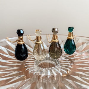 May include: Four jewel-toned perfume bottle pendants with gold chains are displayed on a clear, patterned glass dish. The bottles are black, clear, smoky, and emerald green, each with a faceted stopper.