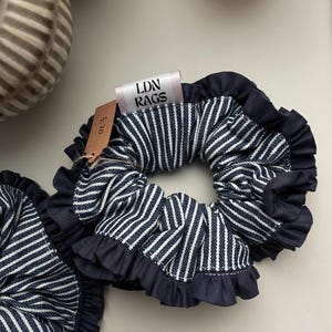 May include: Two navy blue and white striped scrunchies with ruffled edges. The scrunchies have a tag that reads "LDN RAGS". The scrunchies are made of fabric and are designed to hold hair in place.
