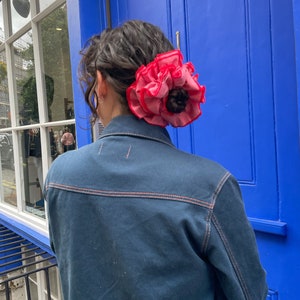 May include: A person with dark brown hair wearing a dark blue denim jacket with a pink and red fabric flower scrunchie in their hair.