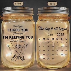 Può includere: Due barattoli di vetro con coperchi argentati, ciascuno con una stringa di luci all'interno. Un barattolo ha il testo "I met you, I liked you, I'm keeping you, forever yours." L'altro ha un calendario per gennaio 2025.