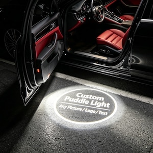 May include: Black car with a red interior and an open door. A custom puddle light shines on the ground, displaying the text "Custom Puddle Light" and "Any Picture/Logo/Text" in white.